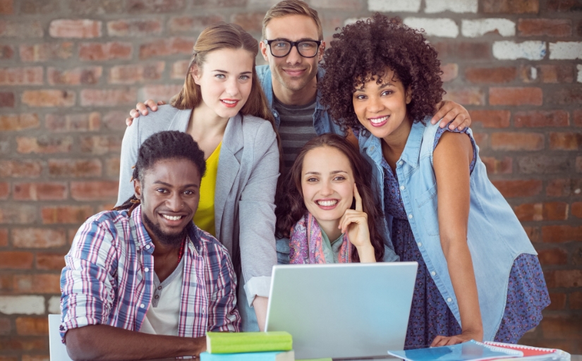Fotografia ilustrativa de cinco pessoas sorrindo para a foto, três mulheres e dois homens. À frente deles existem livros e um notebook.