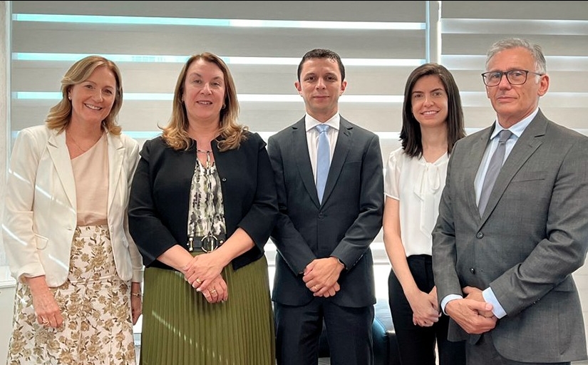 Três mulheres vestidas formalmente e dois homens de terno posam em pé para a foto em uma pequena sala. 