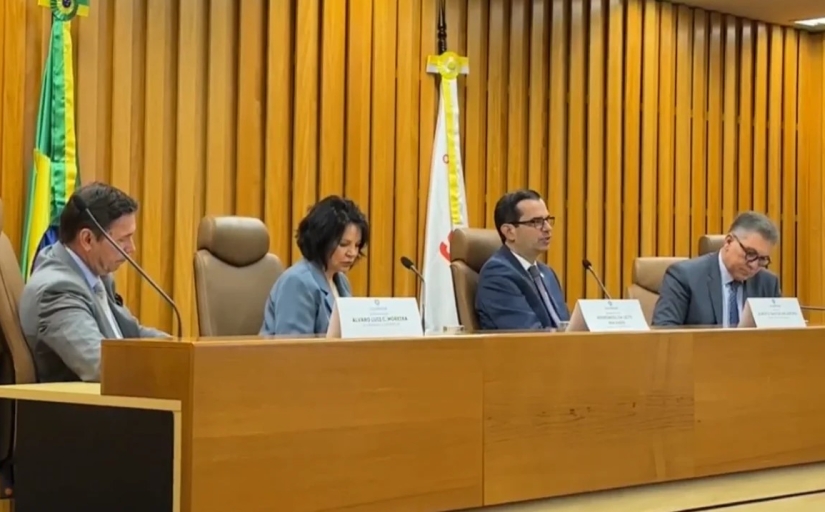 Três homens de terno e uma mulher vestida formalmente sentados em uma mesa de eventos, em tom madeira-ouro, com uma bandeira do Brasil atrás