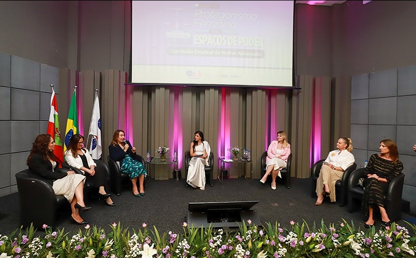 Grupo de sete mulheres sentadas à frente de um auditório, sobre um tablado, formando um semicírculo. Uma delas fala ao microfone