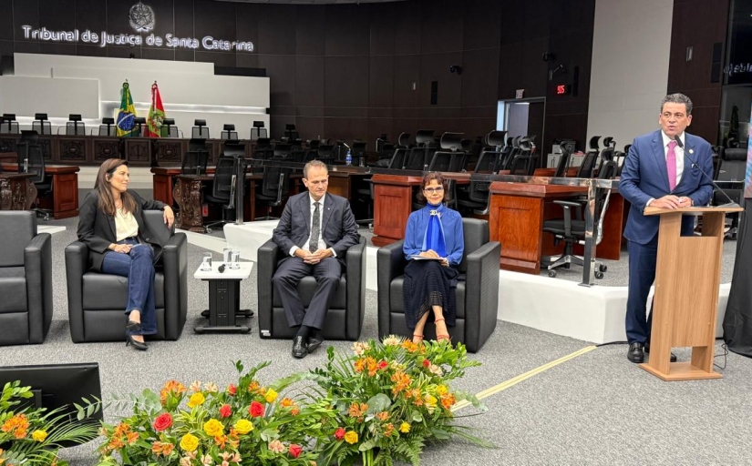 Duas mulheres e um homem sentados em poltronas e um quarto homem, ao lado, falando em um púlpito no auditório do Tribunal de Justiça de SCo