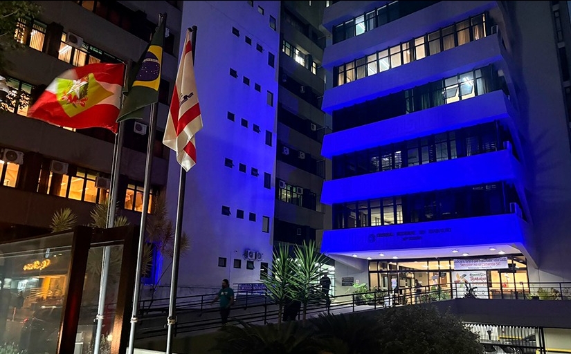 Foto mostra cinco pavimentos do prédio-sede do TRT-SC, iluminado de azul escuro, em ângulo inclinado