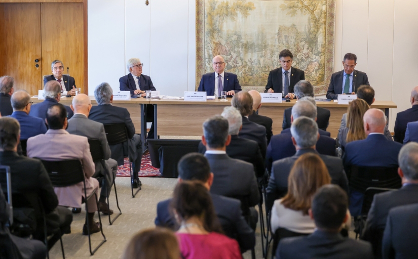 Cinco homens de terno sentados à mesa de um evento observados por outras 21 pessoas em um auditório