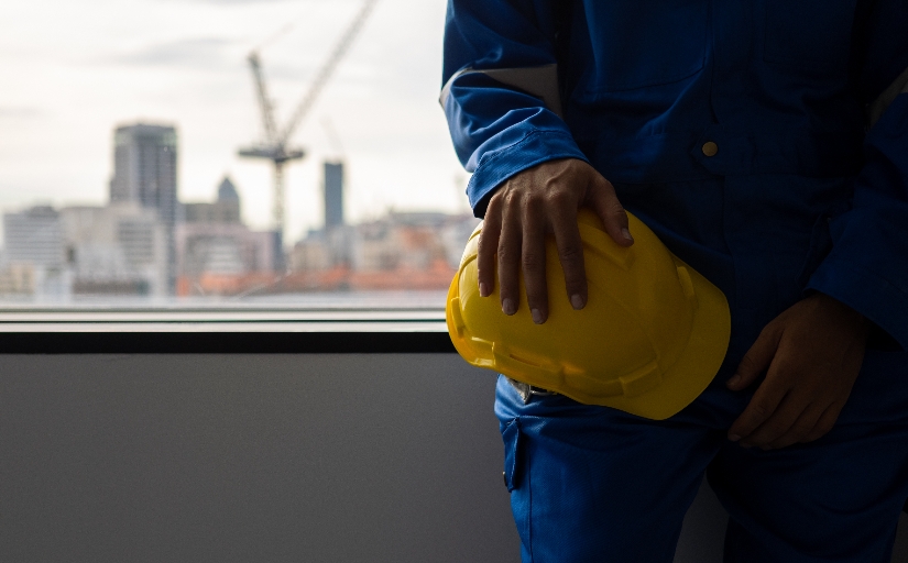 Trabalhador de obra com uniforme azul segura capacete amarelo diante de janela com cidade ao fundo