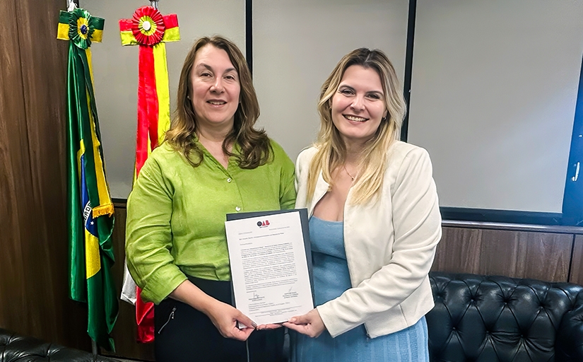 Duas mulheres vestidas formalmente e sorridentes seguram um documento dentro de uma sala