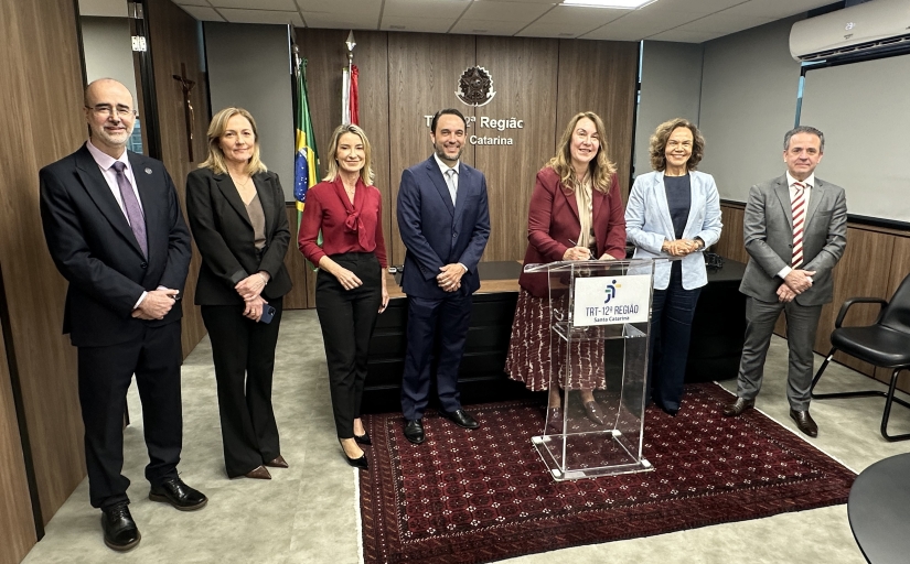 Três homens e quatro mulheres em pé, em uma sala, lado a lado, todos sorridentes. Uma das mulheres assina um documento em um púlpito transparente