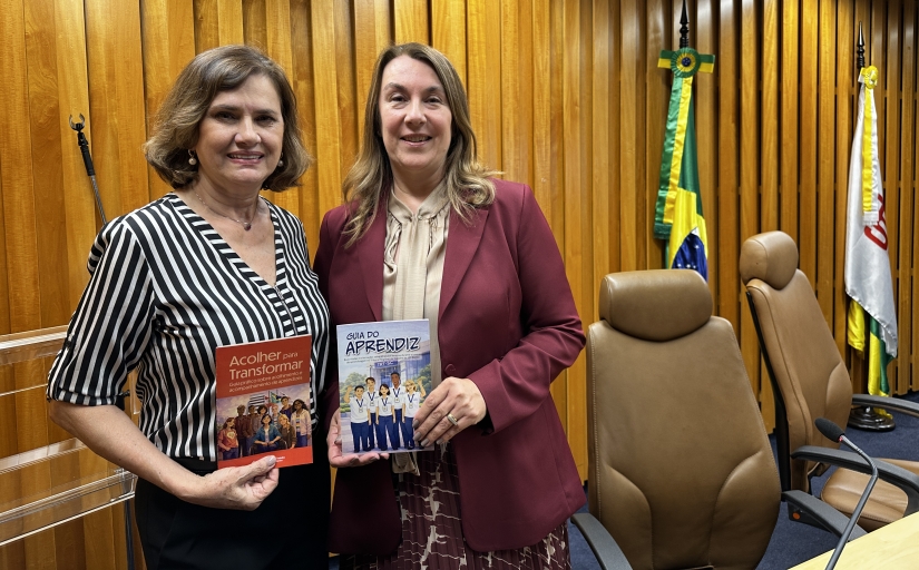 Duas mulheres posam para a foto. Cada uma exibe uma cartilha