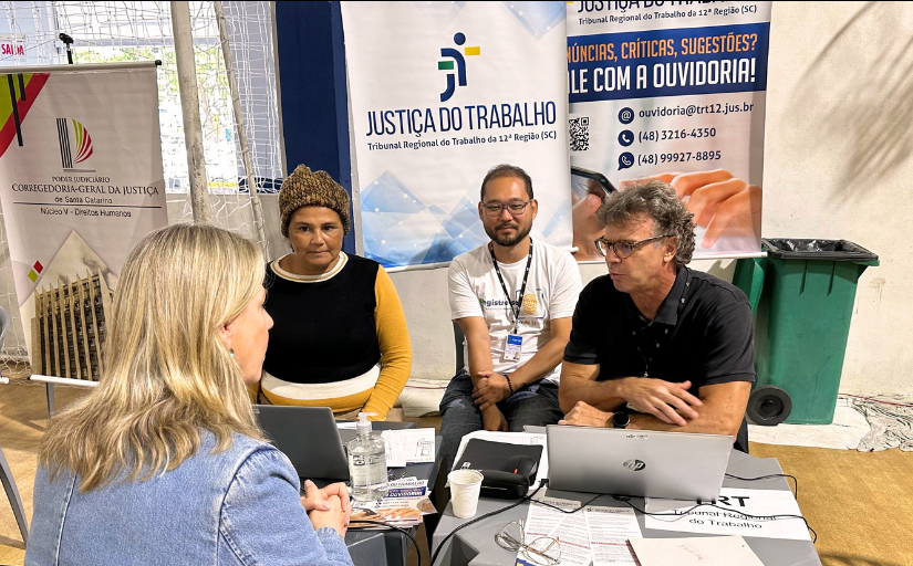 Foto de dois homens e uma mulher prestando atendimento a uma mulher loira. Atrás da mesa, um banner da Justiça do Trabalho