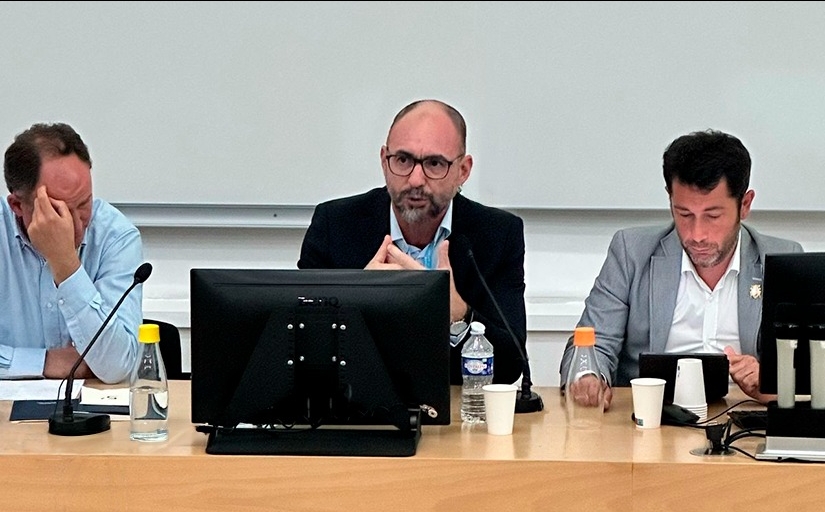 Homem calvo, de óculos, sentado à frente de um notebook, faz uma apresentação. Ao lado dele estão outros dois homens.