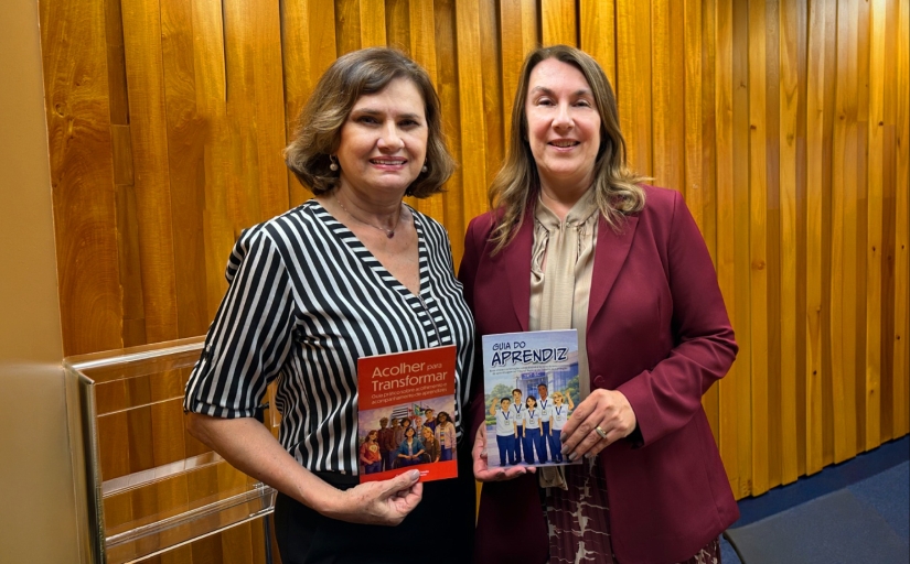Duas mulheres posam para a foto. Cada uma exibe uma cartilha