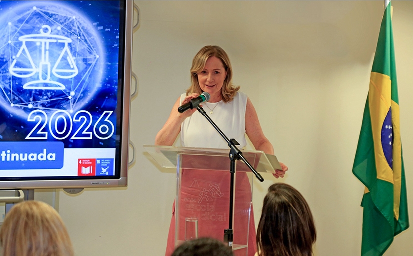 Mulher loira, vestindo blusa branca e saia rosa, fala ao microfone em uma sala de aula. Ela está levemente sorrindo.