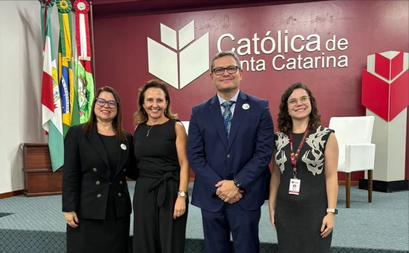 Três mulheres vestidas formalmente e um homem de terno azul marinho posam para a foto, sorridentes, em um auditório. Ao fundo, a logomarca da Universidade Católica