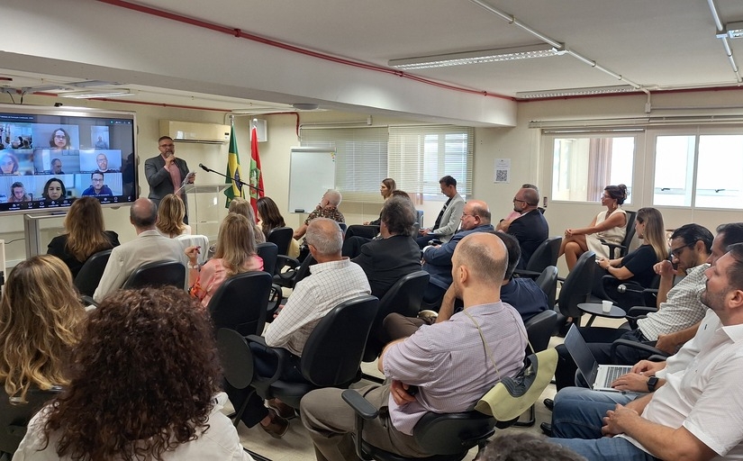 Homem dá palestra a público sentado em sala; bandeiras ao fundo e telão com videoconferência