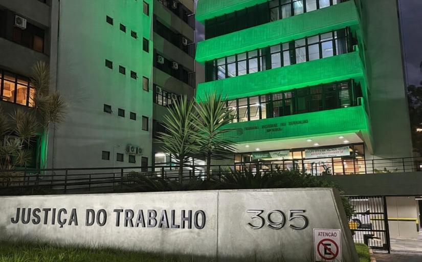 Parte da fachada do TRT-SC iluminada de verde, mostrando três pavimentos. À frente, no chão, um letreiro escrito Justiça do Trabalho