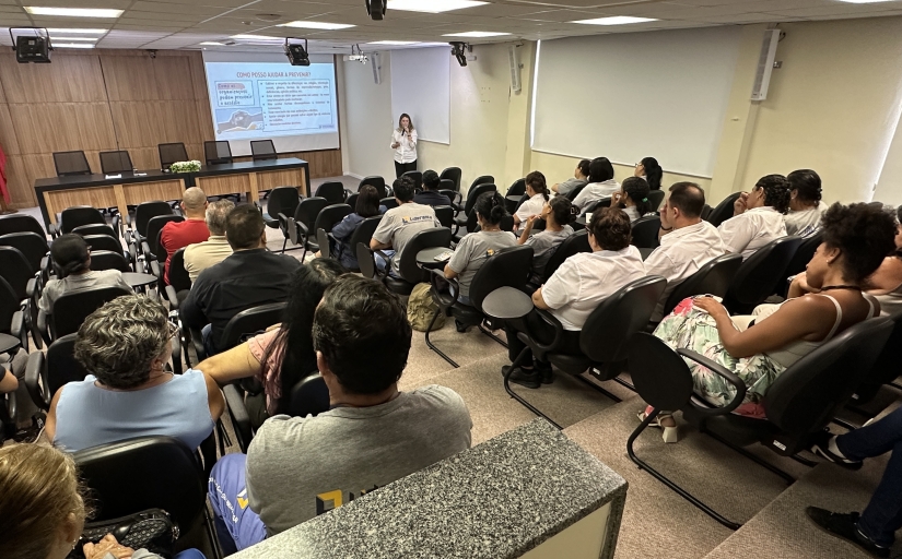 Foto de uma palestra, com pessoas sentadas em um auditório a uma mulher palestrante na frente do público 