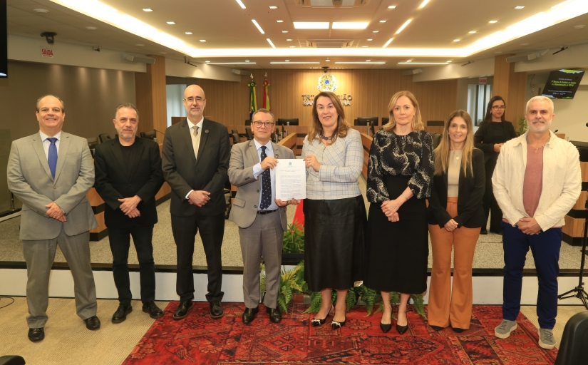 Cinco homens e três mulheres em pé, em uma sala de sessões de julgamento, posam para a foto. Uma mulher e um homem no centro exibem um documento.