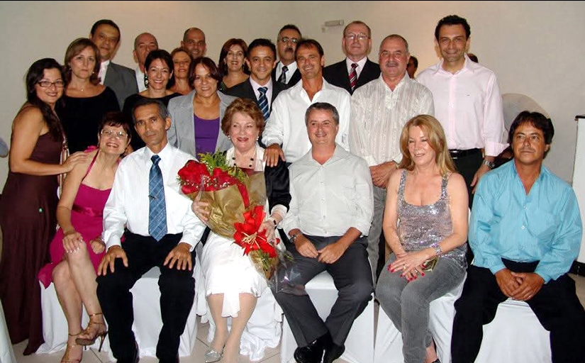 Foto posada de 21 pessoas, algumas sentadas e outras em pé. No centro, Ione Ramos está sentada e segurando, sorridente, uma buque de flores