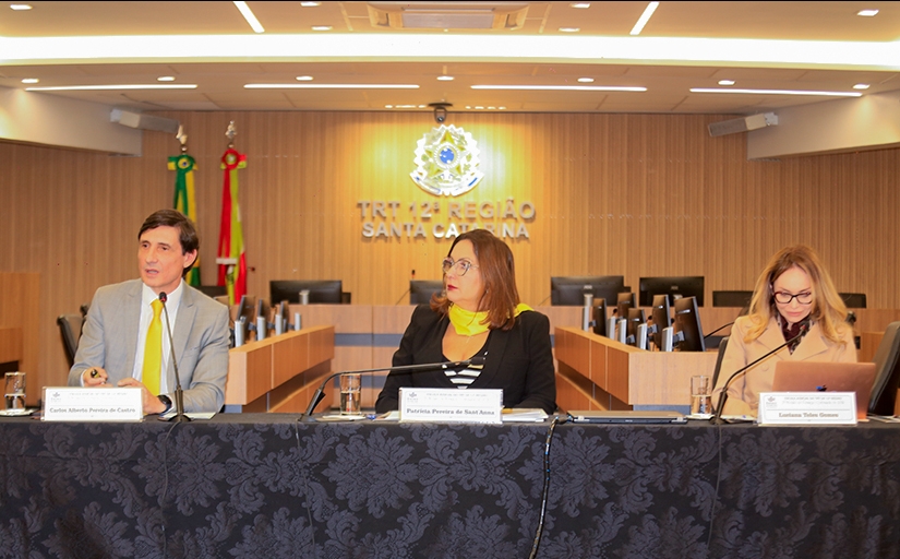 Um homem e duas mulheres estão sentados em uma mesa de honra, durante uma palestra