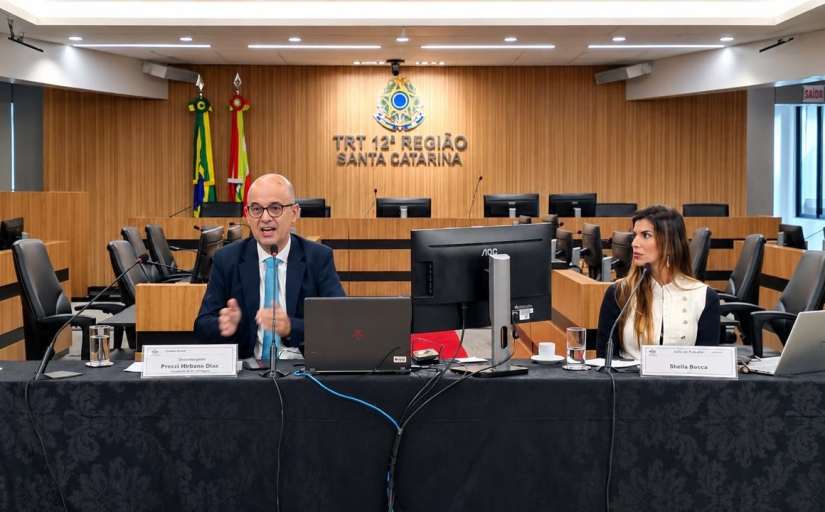 Duas pessoas, um homem e uma mulher, sentados diante de mesa de honra durante palestra em plenário