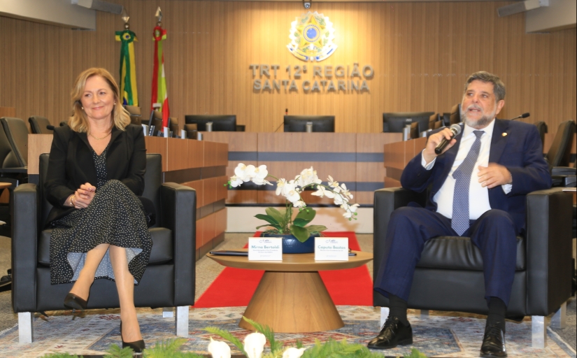 Home de terno escuro fala ao microfone sentado em uma poltrona, na sala de sessões do Pleno do TRT-SC. Ao lado dele, em outra poltrona, uma mulher loira sorridente, com roupa preta