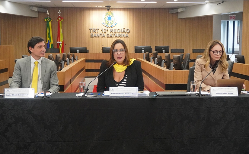 Um homem e duas mulheres estão sentados em uma mesa de honra, durante uma palestra