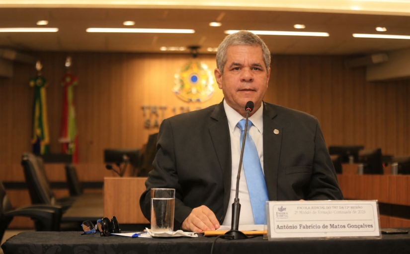 Fotografia de homem sentado falando em uma mesa de honra, durante uma palestra