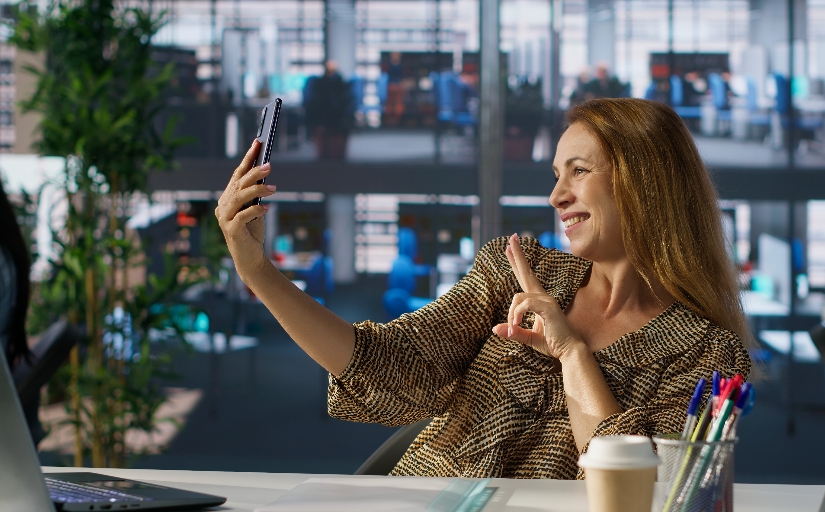 Fotografia ilustrativa de mulher sorrindo para o próprio celular em ambiente de escritório