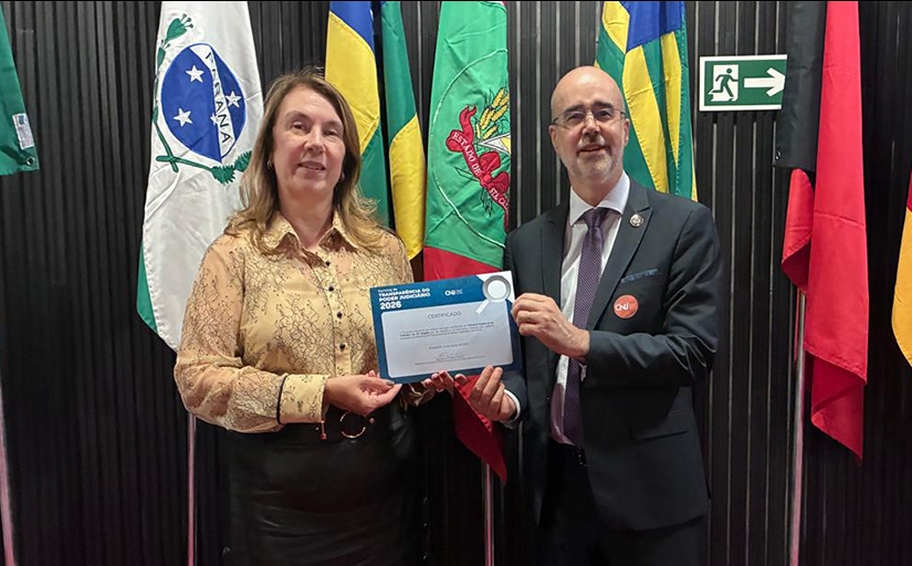 Uma mulher vestida formalmente e um homem de terno e gravata seguram, juntos, um certificado. Ao fundo bandeiras do Brasil e dos estados do Paraná, Santa Catarina e outra não identificada