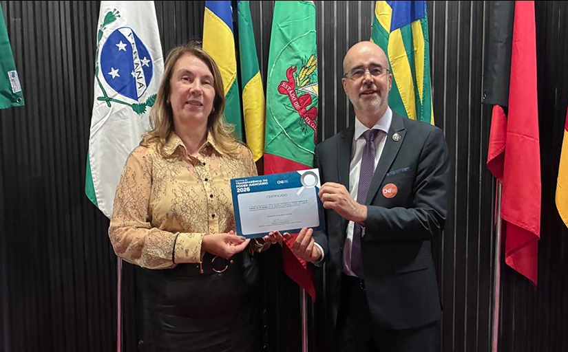 Uma mulher vestida formalmente e um homem de terno e gravata seguram, juntos, um certificado. Ao fundo bandeiras do Brasil e dos estados do Paraná, Santa Catarina e outra não identificada