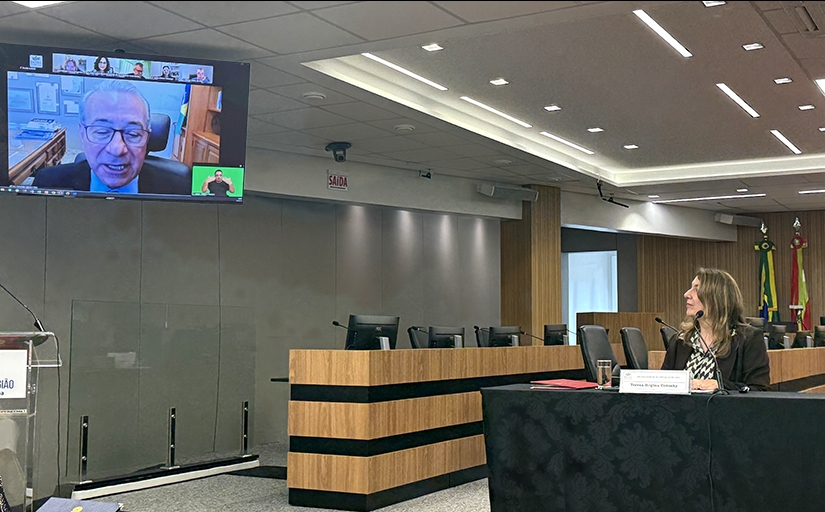Mulher sentada à mesa de um evento observa palestrante que aparece em um telão