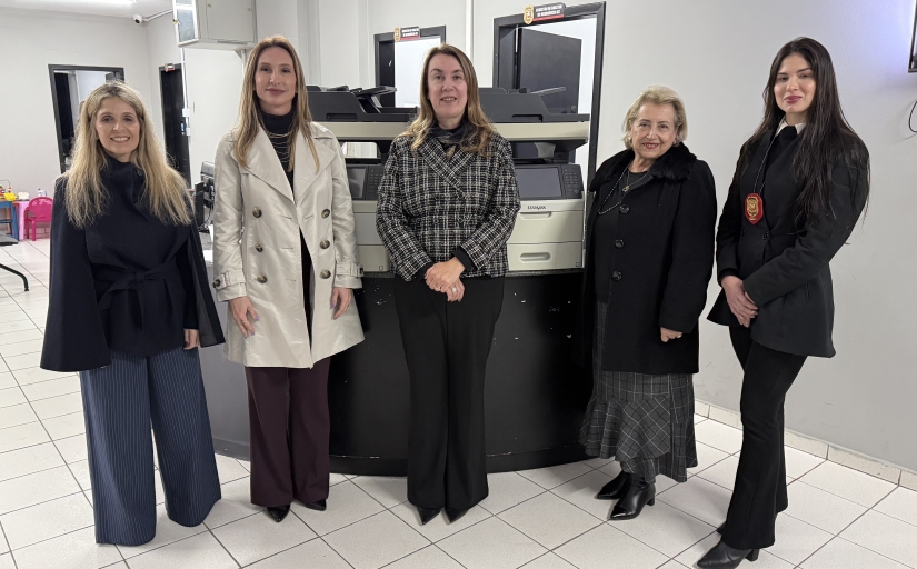 Cinco mulheres posam para a foto. Atrás delas, dois equipamentos de informática