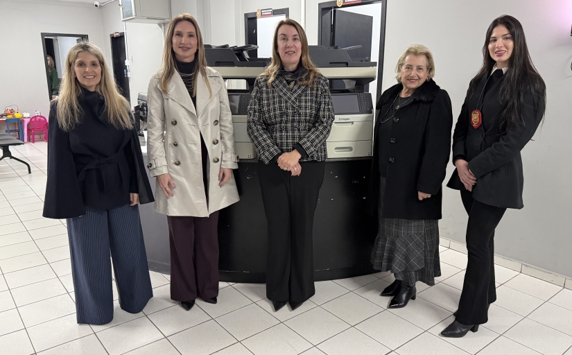 Cinco mulheres posam para a foto. Atrás delas, dois equipamentos de informática