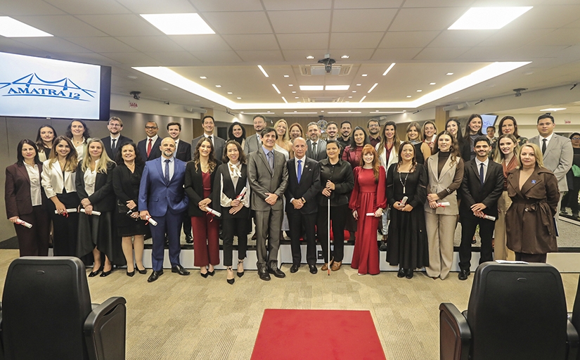 Foto de grupo com cerca de 40 pessoas, entre homens e mulheres, reunidas em um auditório. A maioria está em pé, lado a lado, em frente a uma tela com o logotipo da Amatra 12. As pessoas vestem roupas sociais e algumas seguram certificados. Ao fundo, há cadeiras e equipamentos de apresentação.