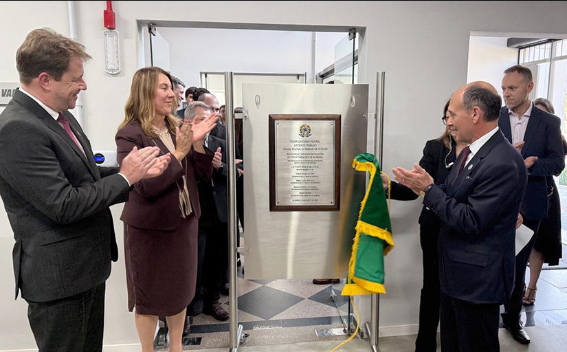 Dois homens de terno e gravata e uma mulher estão aplaudindo felizes em frente a uma placa de inaguração de um forum trabalhista