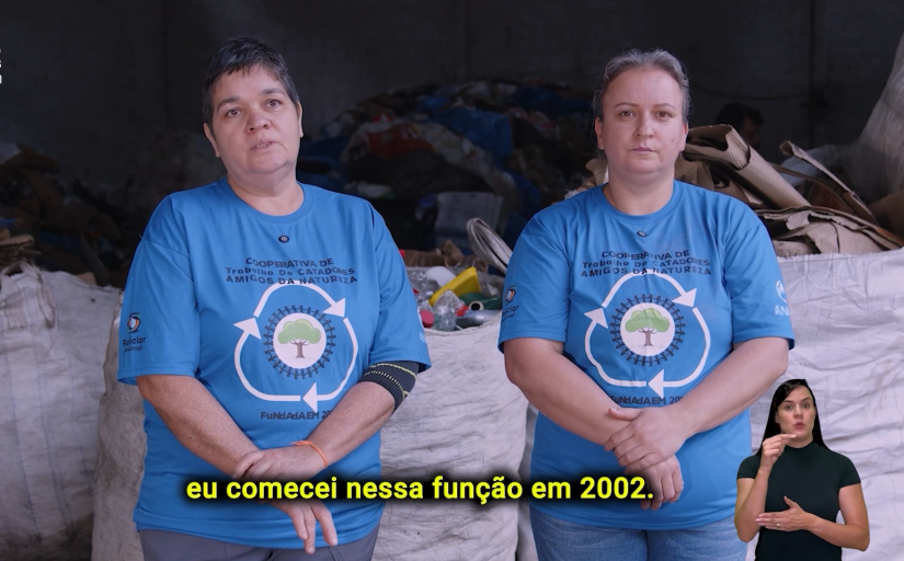 Duas mulheres brancas, uma de cabelos escuros e outra loira, estão lado a lado, olhando para a câmera, à frente de resíduos para reciclagem