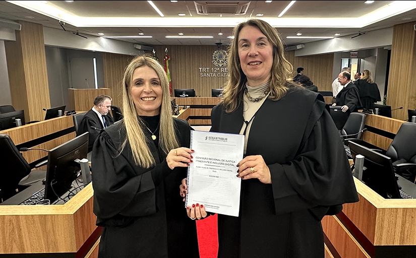Duas mulheres vestindo toga, numa sala de sessão de julgamentos, exibem um documento. Ambas estão sorridentes