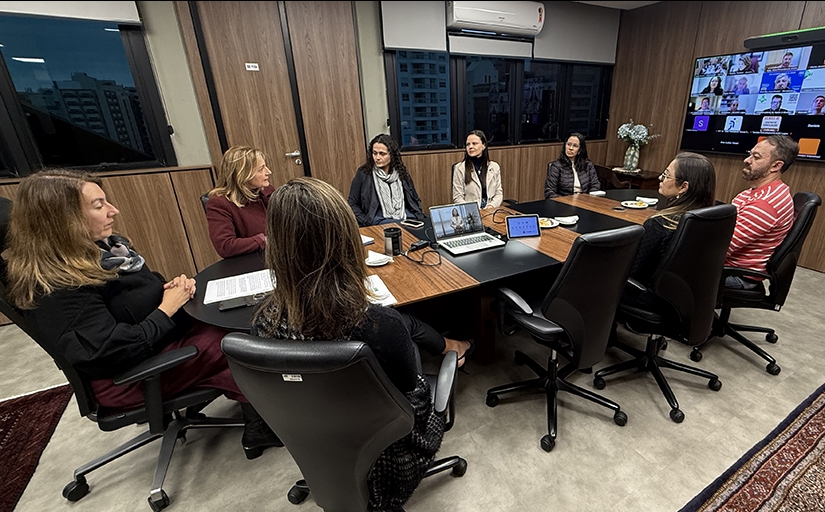 Grupo de sete mulheres e um homem, todos sentados ao redor de uma mesa. Eles observam um telão onde aparecem pessoas em videoconferência