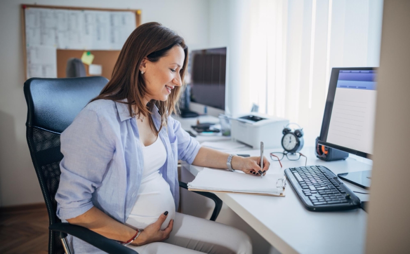 Mulher grávida, sentada, de perfil, trabalhando em um notebook. Ela segura a barriga com um olhar feliz