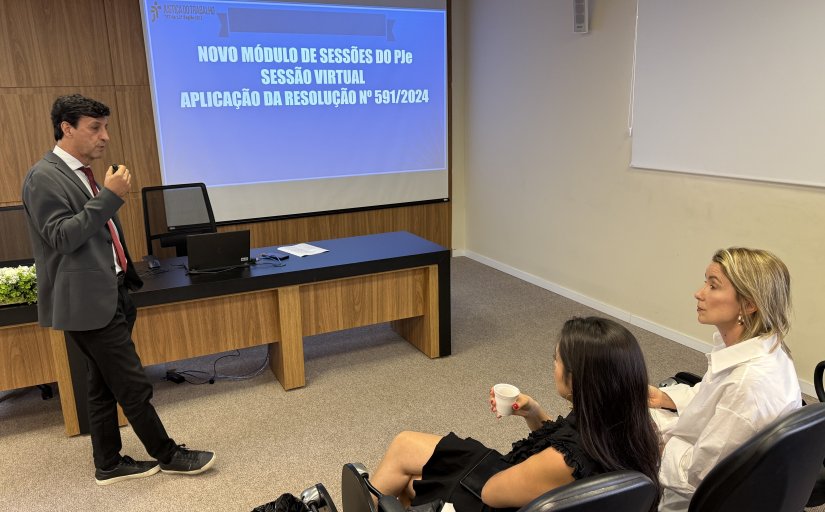 Homem de terno cinza faz uma apresentação a duas mulheres sentadas em um auditório