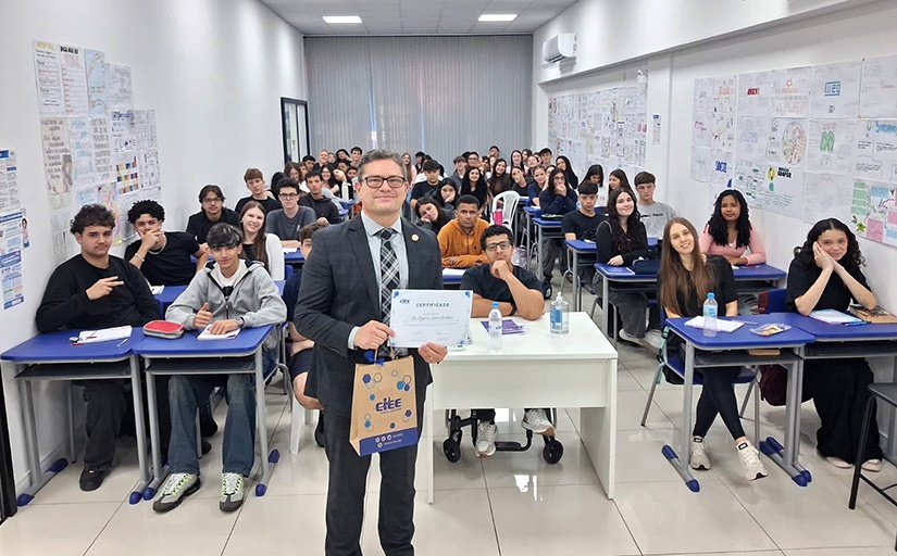 Homem de terno mostrando um certificado em uma sala de aula com diversos adolescentes sentados em carteiras escolares.