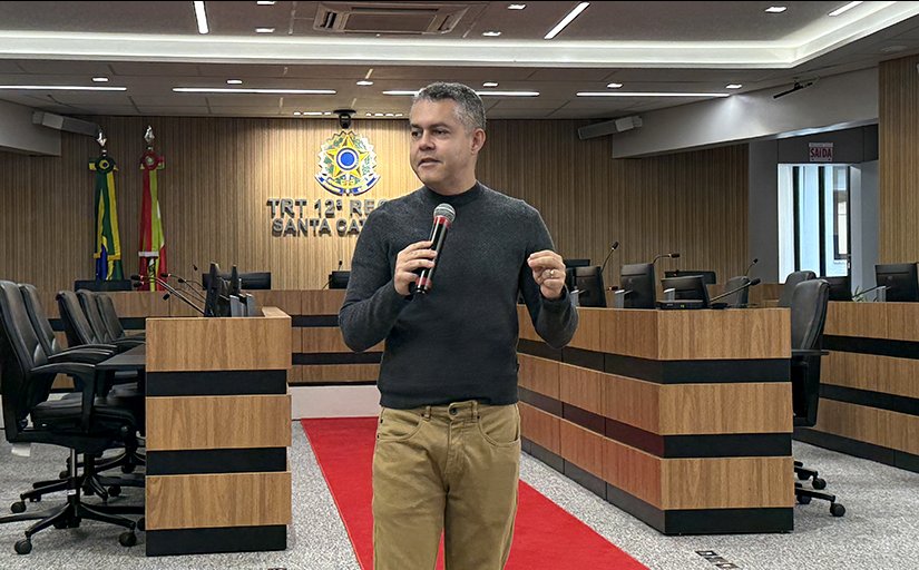 Homem de blusa preta e calça caqui, de pé, fala ao microfone. Ele está em um auditório, tendo ao fundo um brasão do Poder Judiciário iluminado
