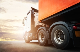 Foto da lateral de um caminhão baú estacionado no asfalto. Ao fundo, ilustração do brilho do sol 