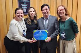 Foto de três mulheres e um homem em pé, posando para a fotografia sorridentes e segurando um certificado azul