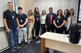 Foto: grupo de três homens e seis mulheres lado a lado, em uma sala, todos sorridentes. À frente, mais à direita, uma mesa de trabalho e dois monitores