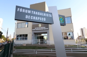 Foto de um prédio claro de dois pavimentos, com rampa de acesso e corrimões. Em primeiro plano, uma placa o identifica, com o letreiro Fórum Trabalhista de Chapecó