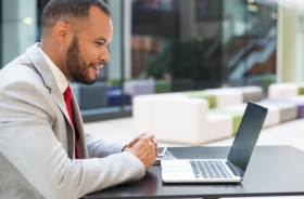 Fotografia ilustrativa de homem negro sorridente vestido formalmente olhando para tela de um notebook ao ar livre