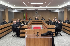 Em um plenário estão sentadas quinze pessoas, sendo três mulheres e doze homens. Eles vestem togas pretas e estão em estações de trabalho posicionadas em quatro fileiras. Ao fundo, estão sentadoes três homens, sendo ao centro, o presidente do TRT-SC.