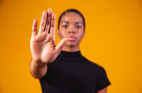 Uma mulher negra está com a mão estendida para frente, com rosto sério e vestida de preto