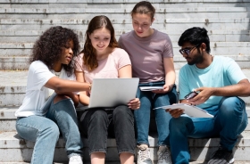 Grupo de quatro estudantes sentados em escadaria ao ar livre, olhando para um notebook com atenção.