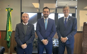 Três homens de terno, lado a lado, posam para foto, dentro de uma sala. À esquerda, aparece a bandeira do Brasil